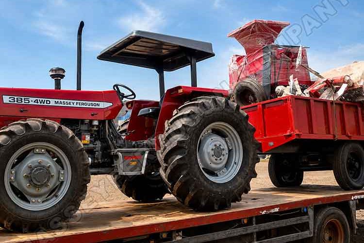 Red Massey Ferguson 385 4WD tractor on a flatbed trailer with a red scrap wagon and piled debris against a blue sky