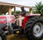 Man in a cap sits on a red Massey Ferguson tractor outdoors with palm trees and a shelter behind him